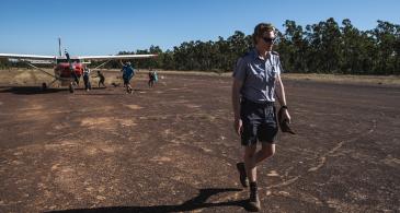 Pilot walking on dirt airstrip with aircraft in background